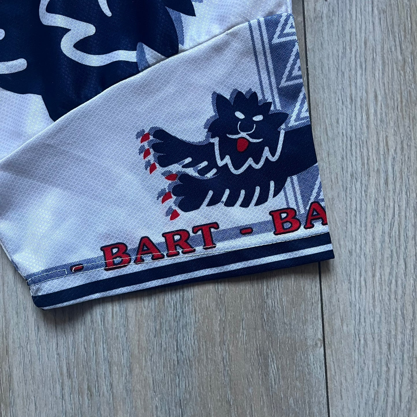 Officially licensed 1999 The Simpsons World Cup T-shirt for England. Lightweight white polyester tee featuring an all-over graphic design. The front shows Bart Simpson kicking a football, dressed in a white tee, with multiple England three-lions emblems layered across the background from top to bottom. Red “Bart” text details appear on the sleeve hems, with Matt Groening / The Simpsons ©1999 licensing printed beneath the main graphic.&nbsp;The back mirrors the artwork in a flipped layout, showing Bart from 