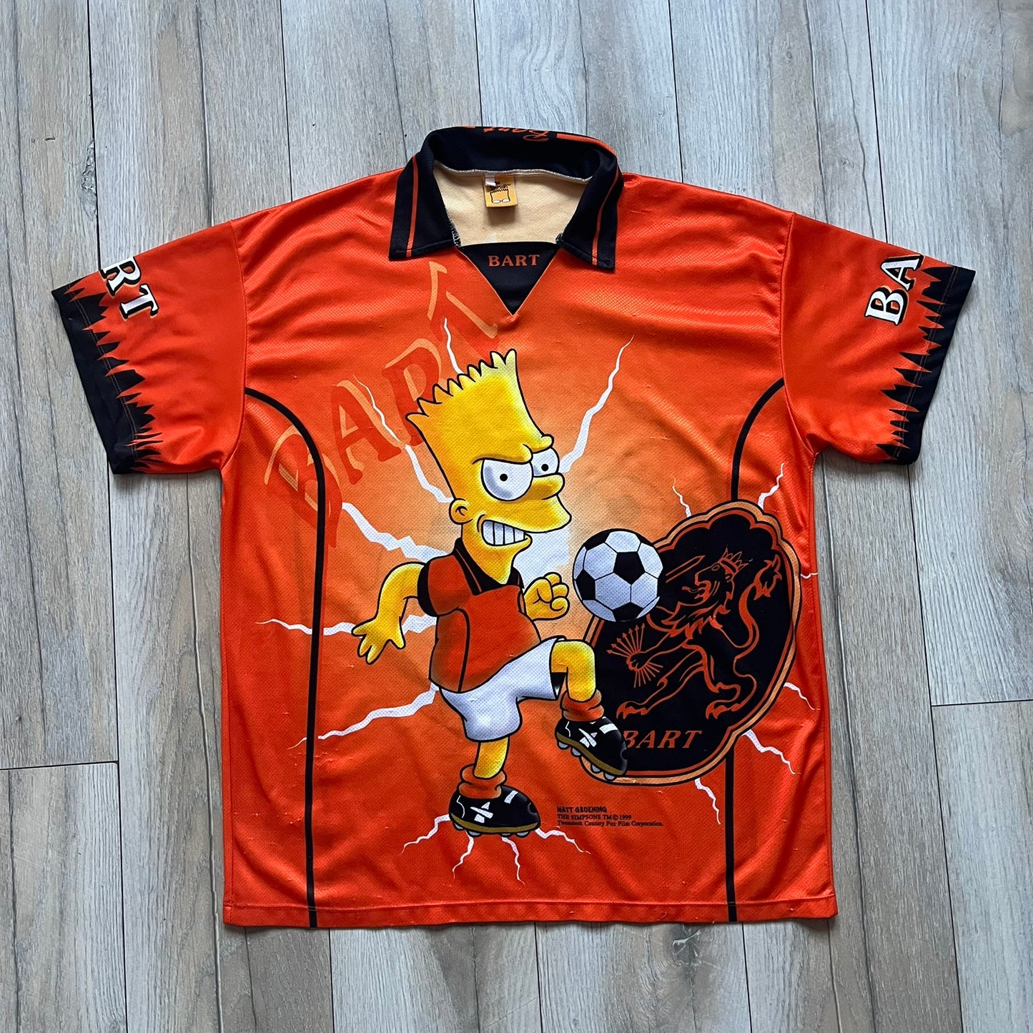 Officially licensed 1999 The Simpsons World Cup Netherlands Tee. Orange lightweight polyester tee with an all-over graphic design. The front features Bart Simpson kicking a football, orange “Bart” text, white lightning graphics, and a Netherlands emblem with Bart illustrated beneath it. Black flame details on the sleeve, along with a black ribbed collar with orange lining. Matt Groening / The Simpsons ©1999 licensing is printed below the main graphic. The back repeats the design in a flipped layout, showing