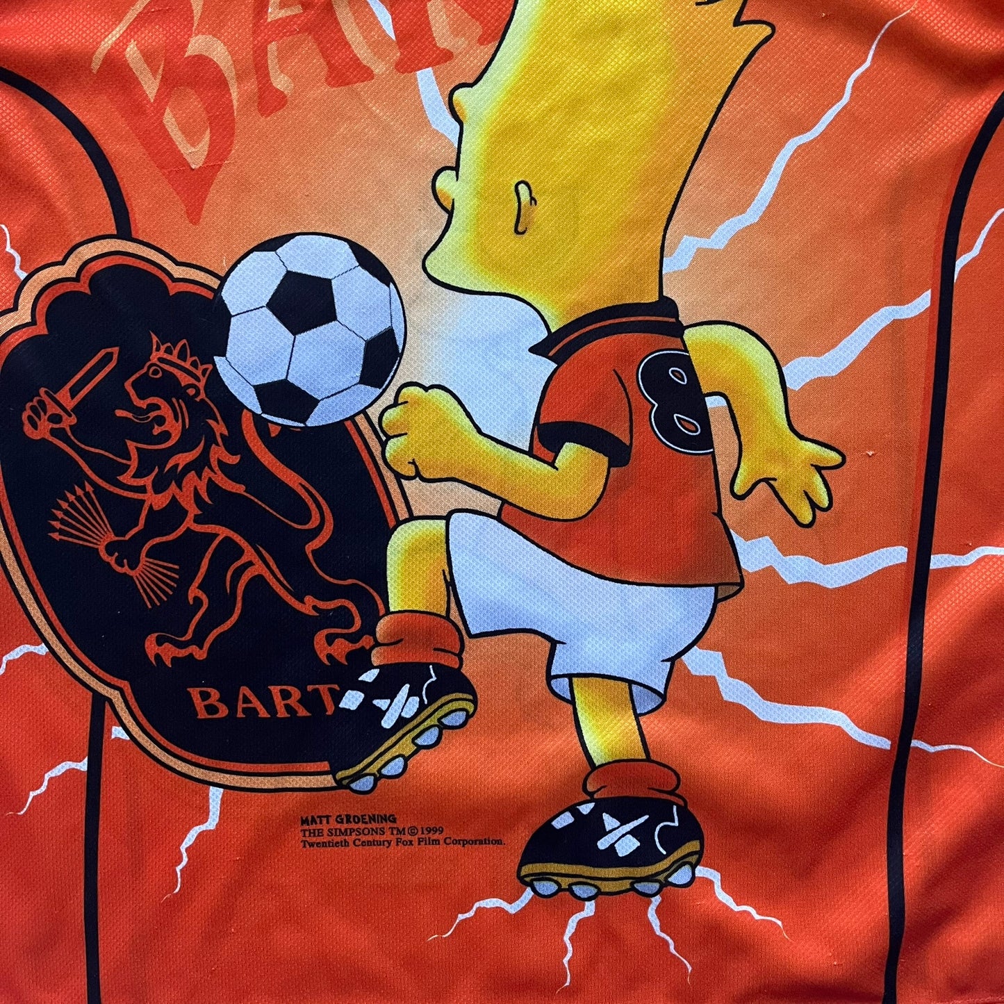 Officially licensed 1999 The Simpsons World Cup Netherlands Tee. Orange lightweight polyester tee with an all-over graphic design. The front features Bart Simpson kicking a football, orange “Bart” text, white lightning graphics, and a Netherlands emblem with Bart illustrated beneath it. Black flame details on the sleeve, along with a black ribbed collar with orange lining. Matt Groening / The Simpsons ©1999 licensing is printed below the main graphic. The back repeats the design in a flipped layout, showing