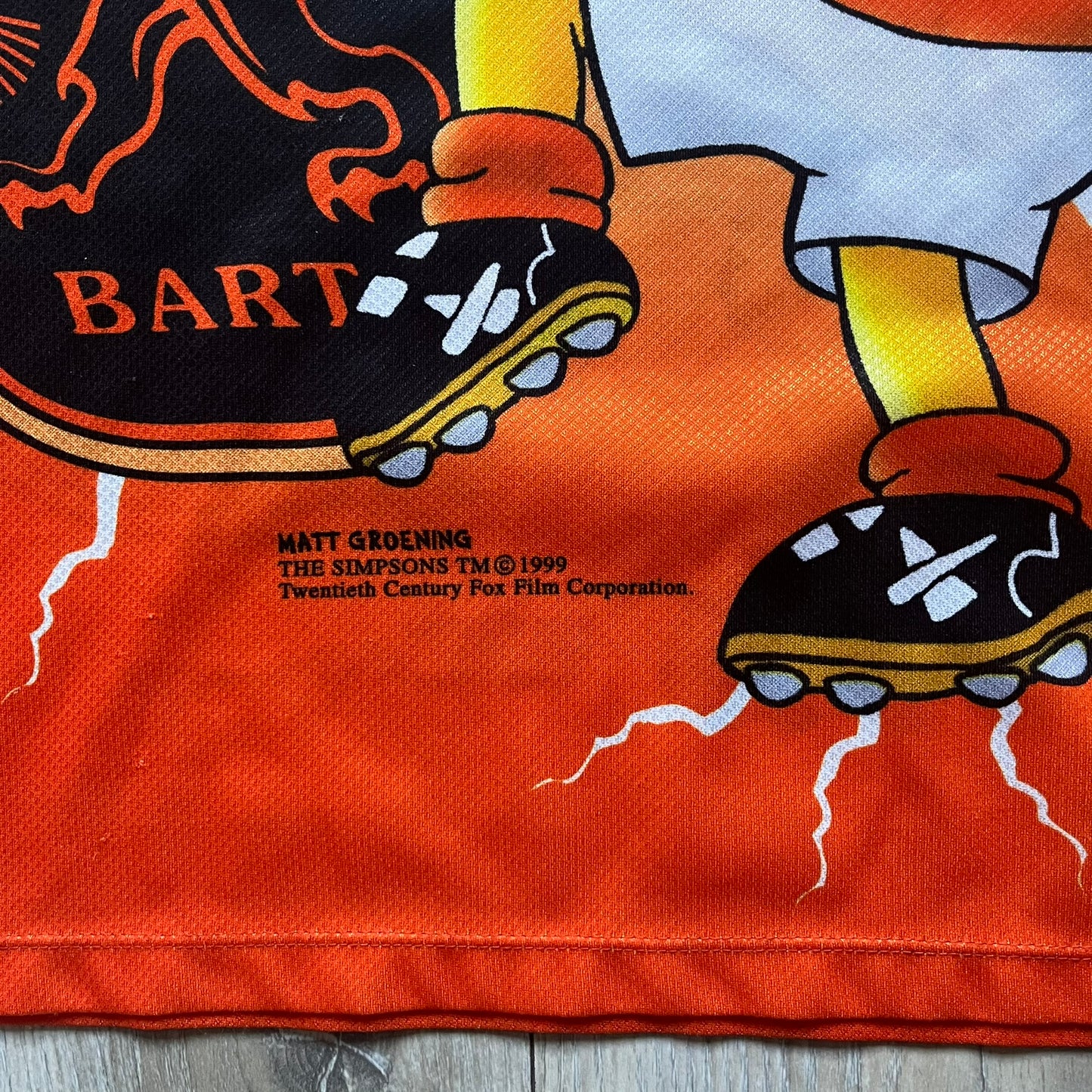 Officially licensed 1999 The Simpsons World Cup Netherlands Tee. Orange lightweight polyester tee with an all-over graphic design. The front features Bart Simpson kicking a football, orange “Bart” text, white lightning graphics, and a Netherlands emblem with Bart illustrated beneath it. Black flame details on the sleeve, along with a black ribbed collar with orange lining. Matt Groening / The Simpsons ©1999 licensing is printed below the main graphic. The back repeats the design in a flipped layout, showing