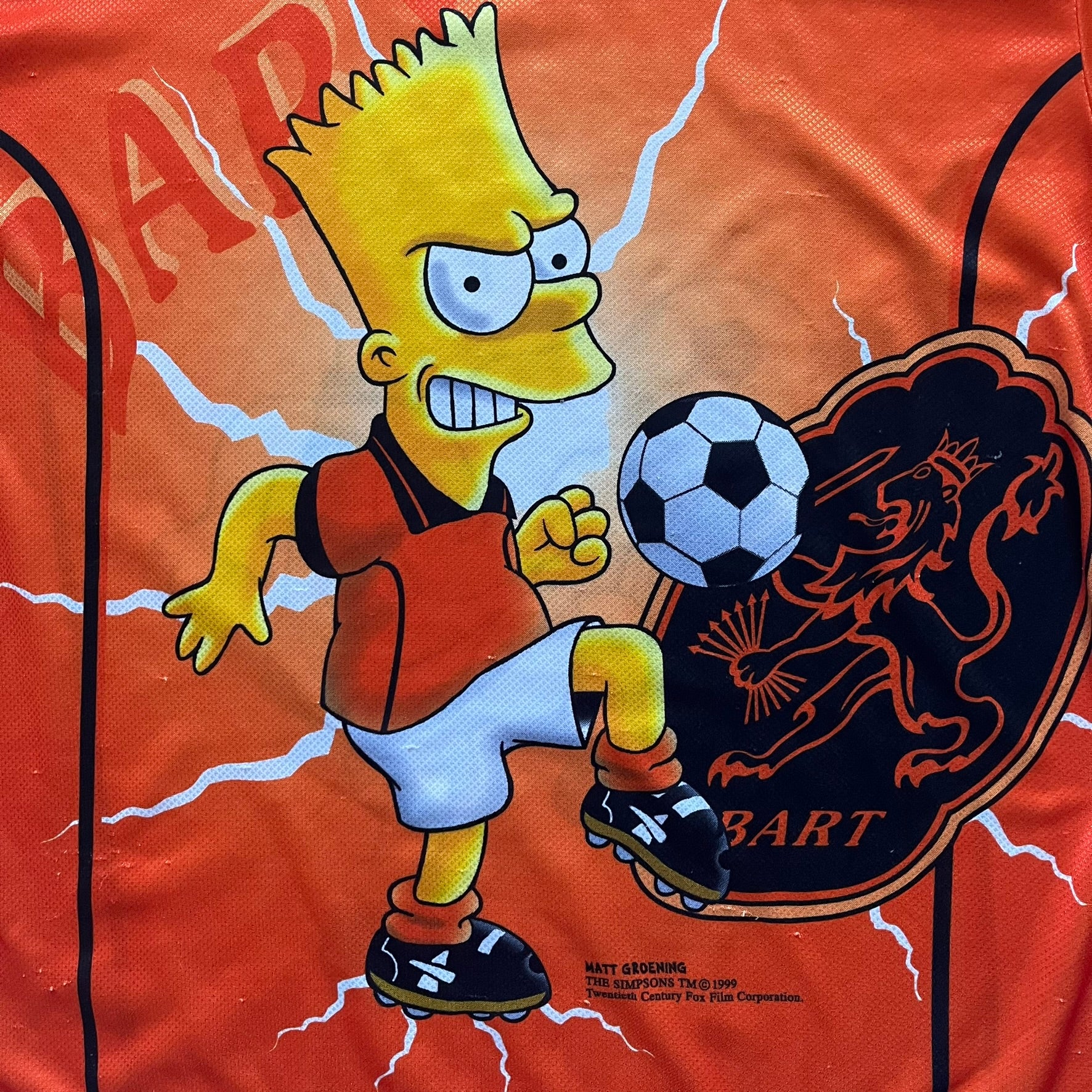 Officially licensed 1999 The Simpsons World Cup Netherlands Tee. Orange lightweight polyester tee with an all-over graphic design. The front features Bart Simpson kicking a football, orange “Bart” text, white lightning graphics, and a Netherlands emblem with Bart illustrated beneath it. Black flame details on the sleeve, along with a black ribbed collar with orange lining. Matt Groening / The Simpsons ©1999 licensing is printed below the main graphic. The back repeats the design in a flipped layout, showing