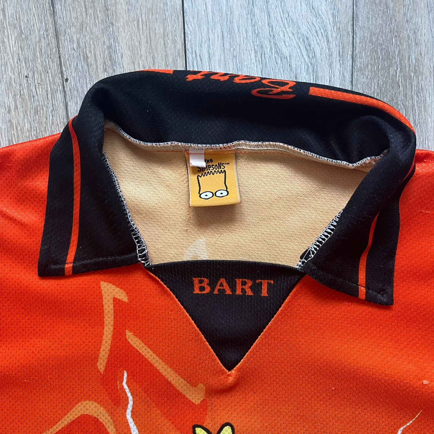 Officially licensed 1999 The Simpsons World Cup Netherlands Tee. Orange lightweight polyester tee with an all-over graphic design. The front features Bart Simpson kicking a football, orange “Bart” text, white lightning graphics, and a Netherlands emblem with Bart illustrated beneath it. Black flame details on the sleeve, along with a black ribbed collar with orange lining. Matt Groening / The Simpsons ©1999 licensing is printed below the main graphic. The back repeats the design in a flipped layout, showing