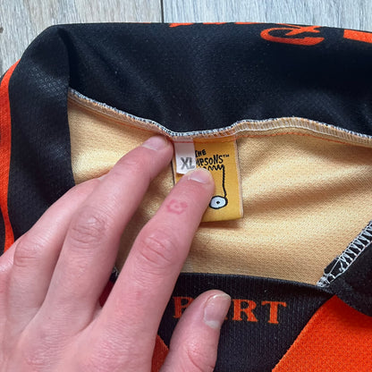 Officially licensed 1999 The Simpsons World Cup Netherlands Tee. Orange lightweight polyester tee with an all-over graphic design. The front features Bart Simpson kicking a football, orange “Bart” text, white lightning graphics, and a Netherlands emblem with Bart illustrated beneath it. Black flame details on the sleeve, along with a black ribbed collar with orange lining. Matt Groening / The Simpsons ©1999 licensing is printed below the main graphic. The back repeats the design in a flipped layout, showing