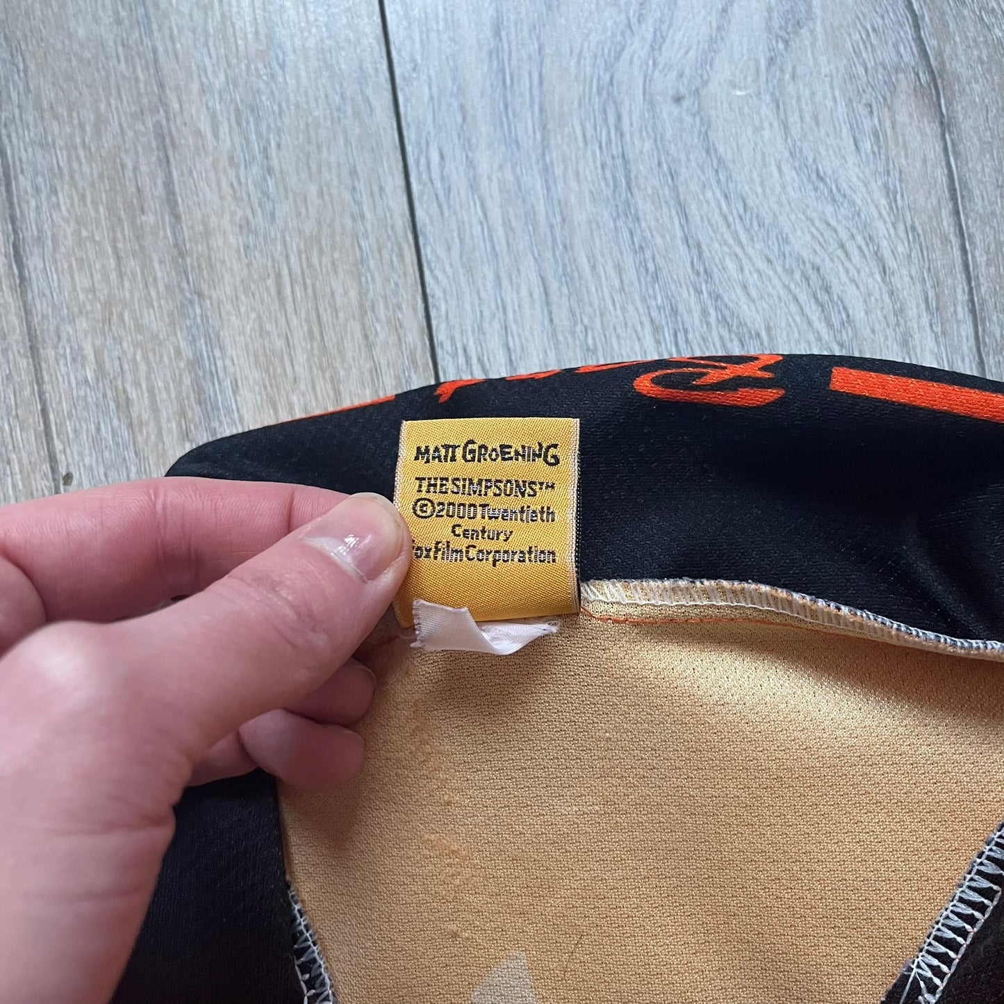 Officially licensed 1999 The Simpsons World Cup Netherlands Tee. Orange lightweight polyester tee with an all-over graphic design. The front features Bart Simpson kicking a football, orange “Bart” text, white lightning graphics, and a Netherlands emblem with Bart illustrated beneath it. Black flame details on the sleeve, along with a black ribbed collar with orange lining. Matt Groening / The Simpsons ©1999 licensing is printed below the main graphic. The back repeats the design in a flipped layout, showing