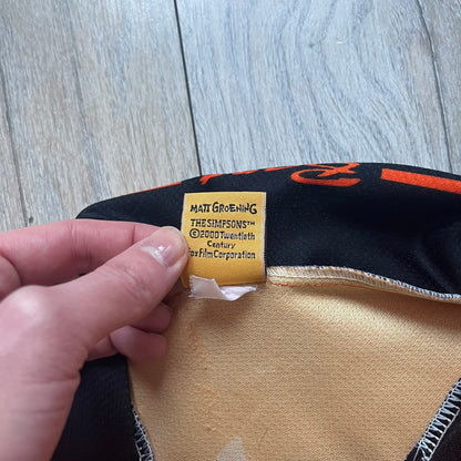 Officially licensed 1999 The Simpsons World Cup Netherlands Tee. Orange lightweight polyester tee with an all-over graphic design. The front features Bart Simpson kicking a football, orange “Bart” text, white lightning graphics, and a Netherlands emblem with Bart illustrated beneath it. Black flame details on the sleeve, along with a black ribbed collar with orange lining. Matt Groening / The Simpsons ©1999 licensing is printed below the main graphic. The back repeats the design in a flipped layout, showing