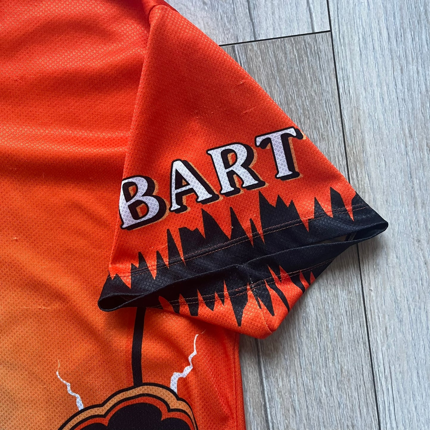 Officially licensed 1999 The Simpsons World Cup Netherlands Tee. Orange lightweight polyester tee with an all-over graphic design. The front features Bart Simpson kicking a football, orange “Bart” text, white lightning graphics, and a Netherlands emblem with Bart illustrated beneath it. Black flame details on the sleeve, along with a black ribbed collar with orange lining. Matt Groening / The Simpsons ©1999 licensing is printed below the main graphic. The back repeats the design in a flipped layout, showing