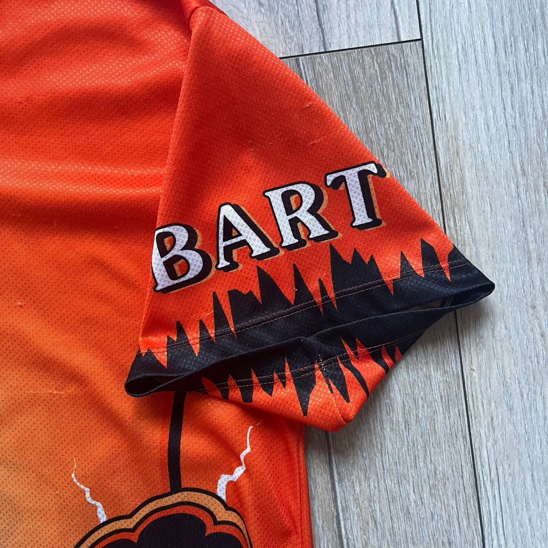 Officially licensed 1999 The Simpsons World Cup Netherlands Tee. Orange lightweight polyester tee with an all-over graphic design. The front features Bart Simpson kicking a football, orange “Bart” text, white lightning graphics, and a Netherlands emblem with Bart illustrated beneath it. Black flame details on the sleeve, along with a black ribbed collar with orange lining. Matt Groening / The Simpsons ©1999 licensing is printed below the main graphic. The back repeats the design in a flipped layout, showing