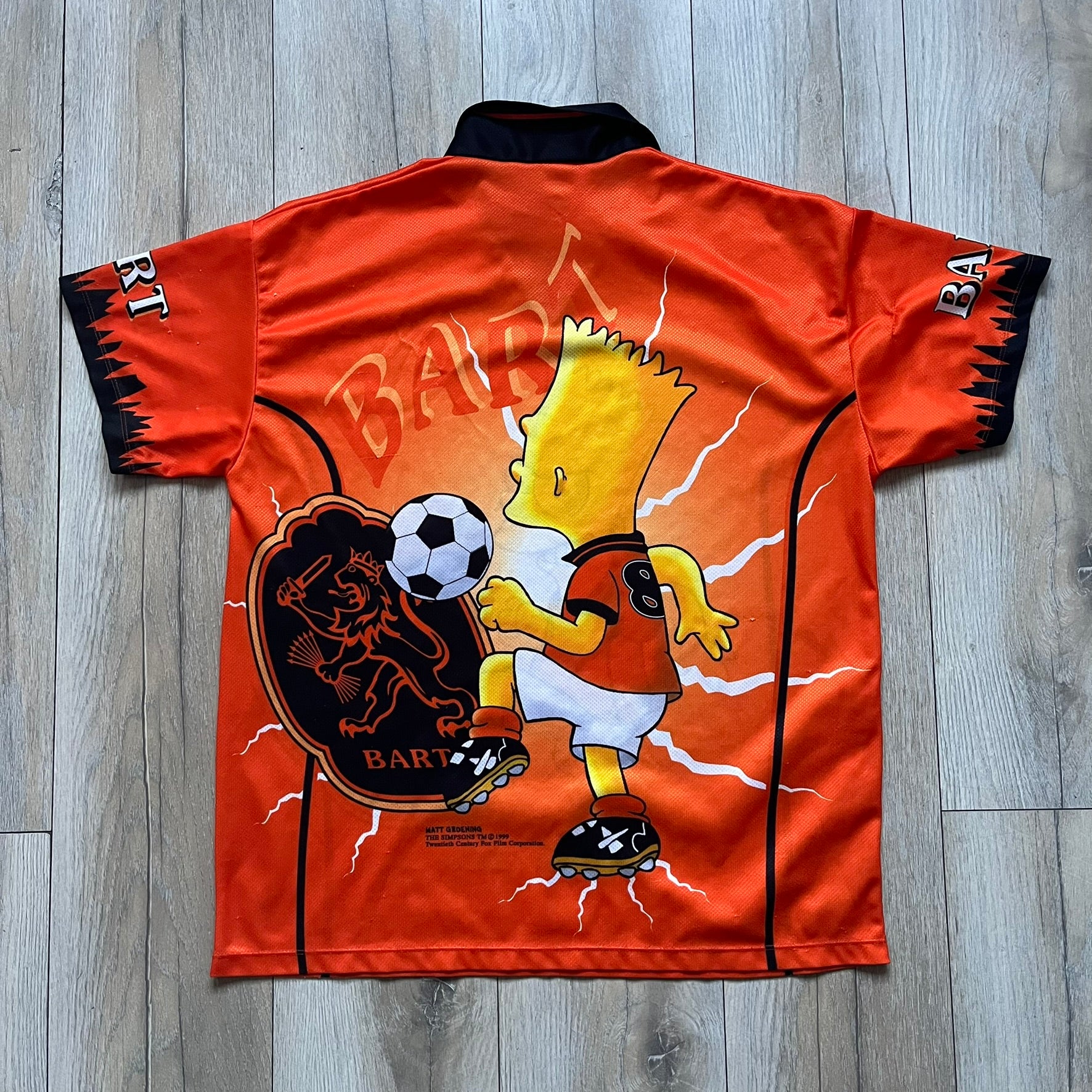 Officially licensed 1999 The Simpsons World Cup Netherlands Tee. Orange lightweight polyester tee with an all-over graphic design. The front features Bart Simpson kicking a football, orange “Bart” text, white lightning graphics, and a Netherlands emblem with Bart illustrated beneath it. Black flame details on the sleeve, along with a black ribbed collar with orange lining. Matt Groening / The Simpsons ©1999 licensing is printed below the main graphic. The back repeats the design in a flipped layout, showing