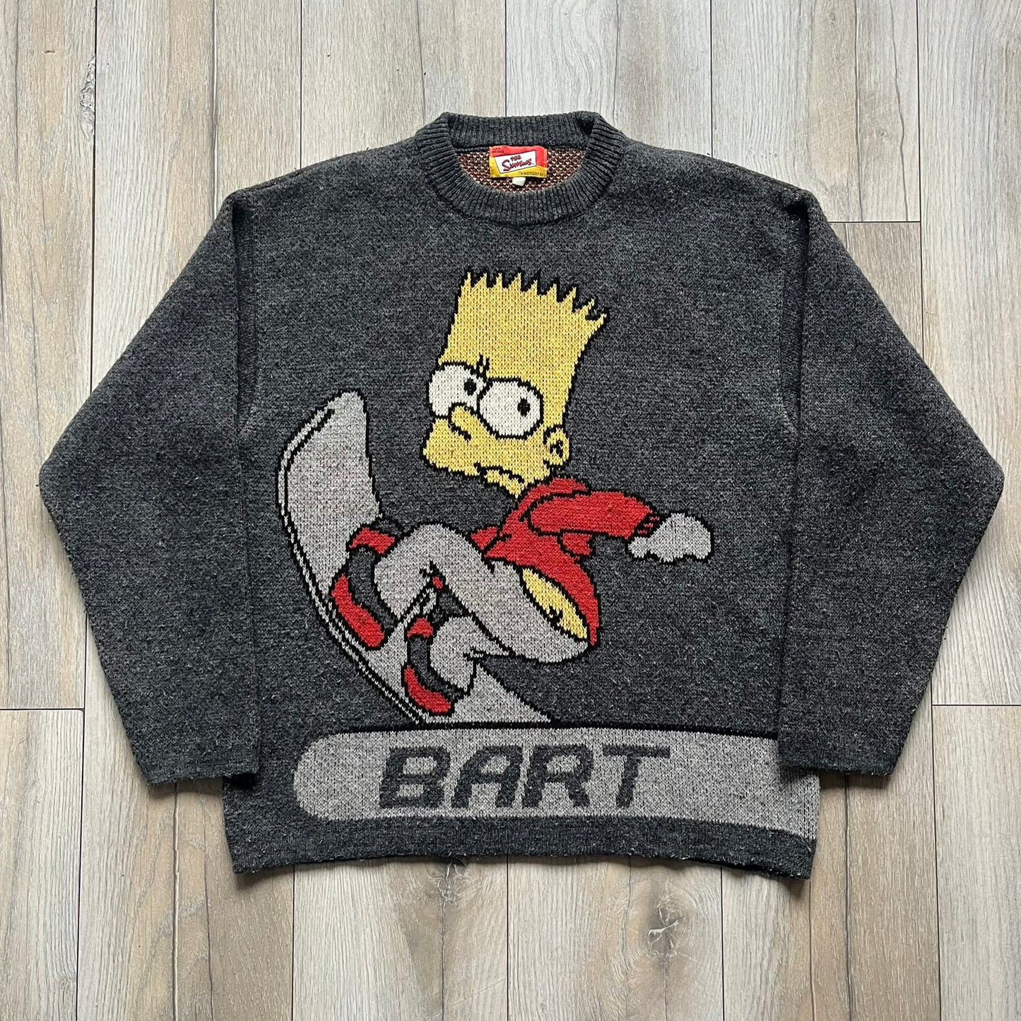 The Simpsons “Bart Freestyle” 2003 Knit Sweater is a gray knit featuring a large woven graphic of Bart snowboarding on the front, with “Bart” text placed below the character. On the back, in the same position as the front graphic, there is “Freestyle” text.&nbsp;It includes a Matt Groening The Simpsons 2003 FOX neck tag.