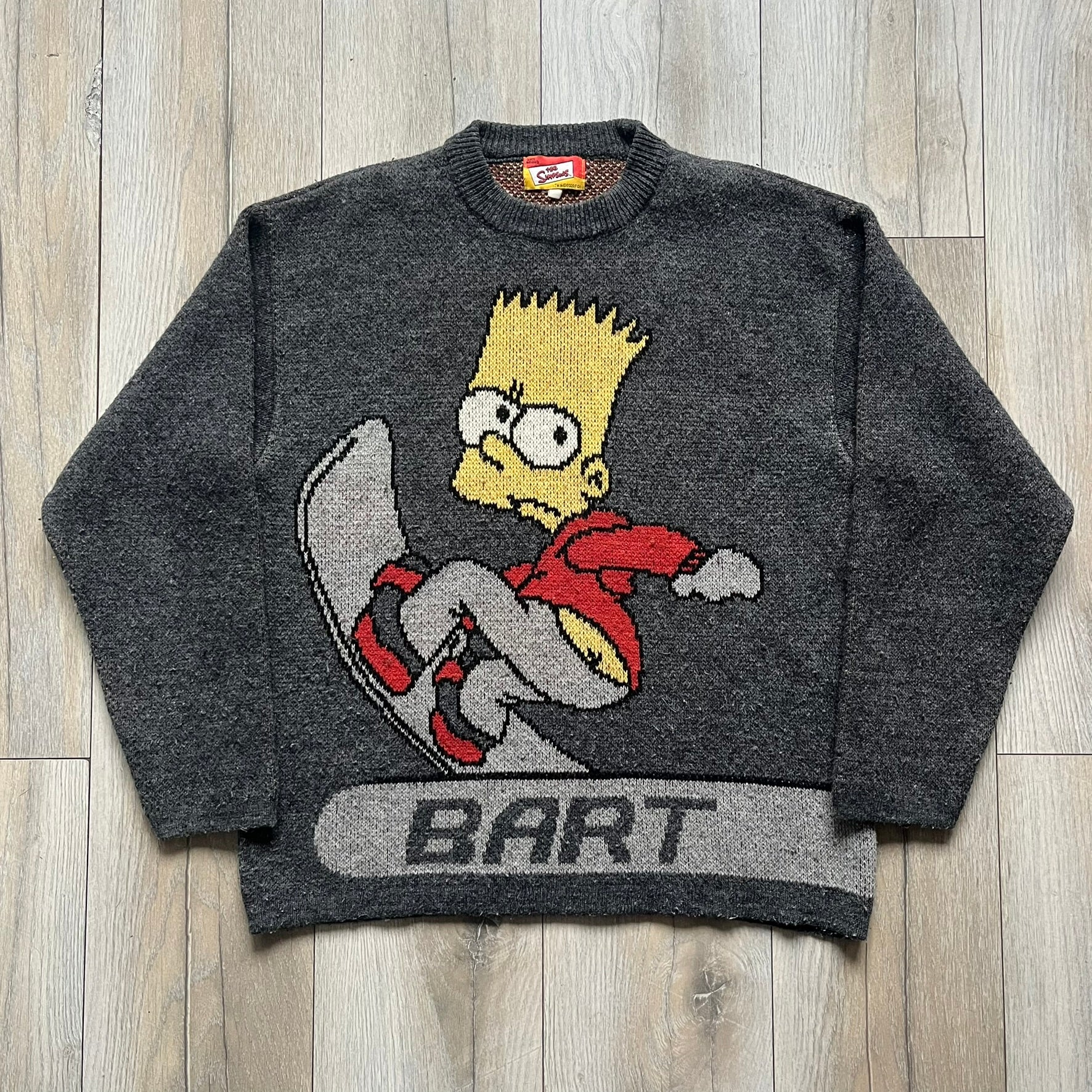 The Simpsons “Bart Freestyle” 2003 Knit Sweater is a gray knit featuring a large woven graphic of Bart snowboarding on the front, with “Bart” text placed below the character. On the back, in the same position as the front graphic, there is “Freestyle” text.&nbsp;It includes a Matt Groening The Simpsons 2003 FOX neck tag.