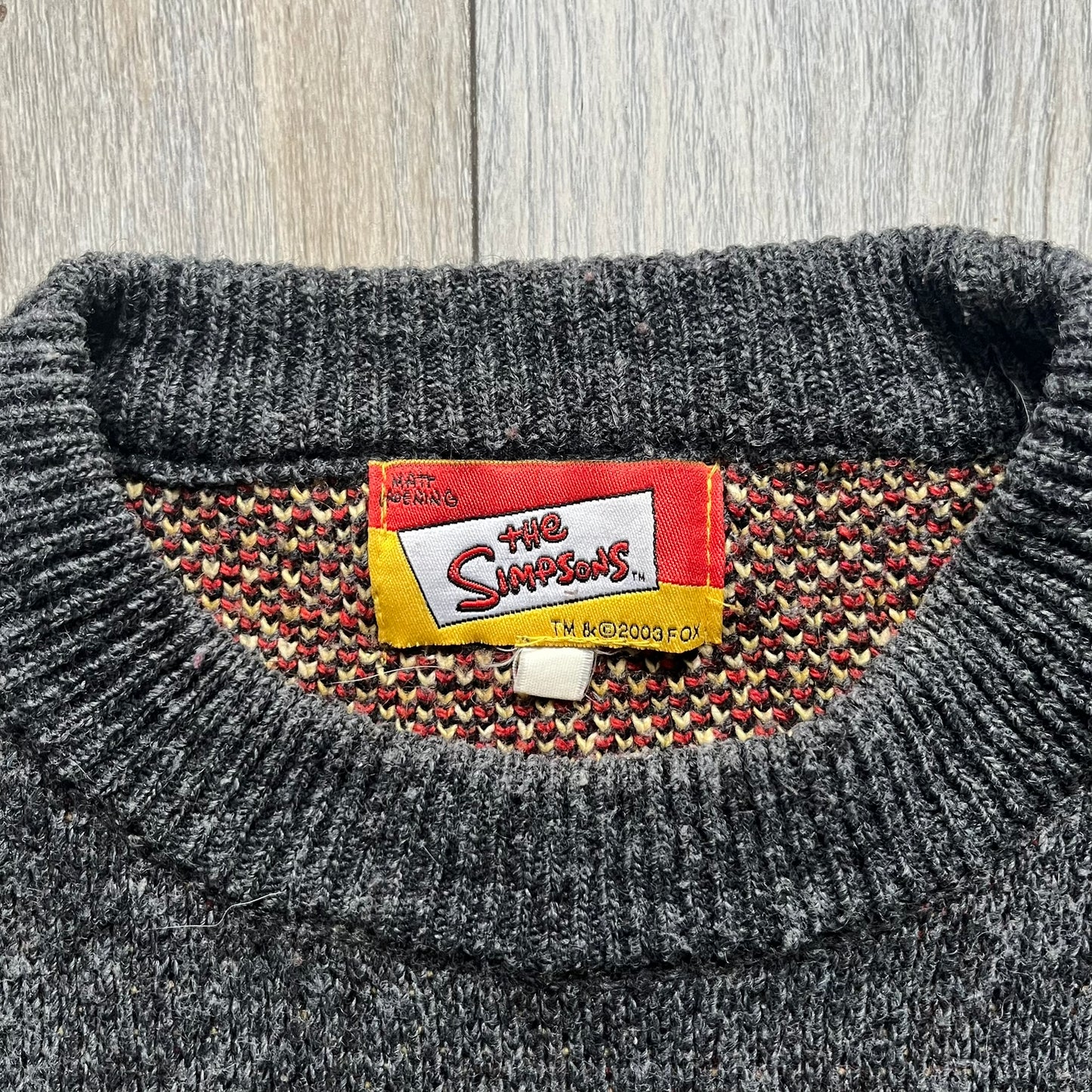 The Simpsons “Bart Freestyle” 2003 Knit Sweater is a gray knit featuring a large woven graphic of Bart snowboarding on the front, with “Bart” text placed below the character. On the back, in the same position as the front graphic, there is “Freestyle” text.&nbsp;It includes a Matt Groening The Simpsons 2003 FOX neck tag.