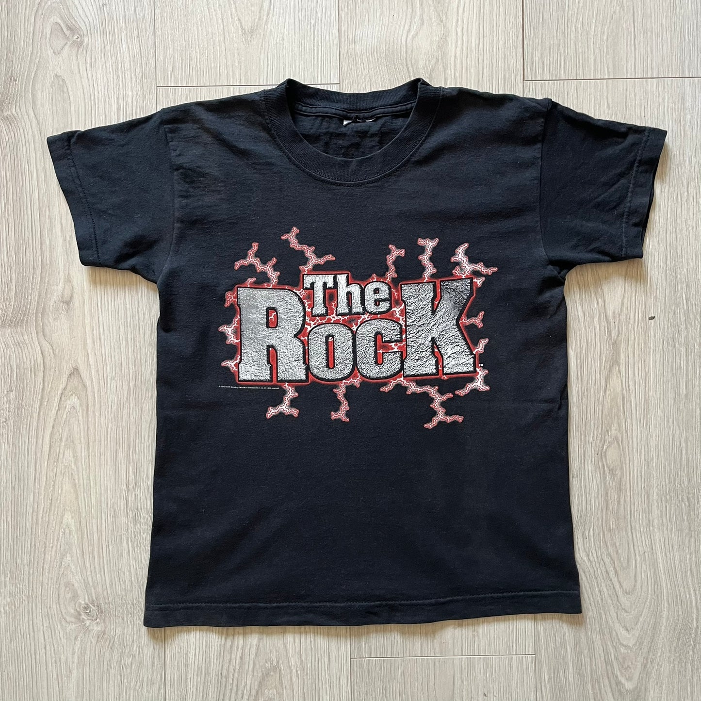 The Rock “Most Electrifying Man” 2000 Tee is a vintage WWE shirt featuring bold metallic-textured lettering with red lightning accents. The front proudly displays “The Rock,” while the back proclaims “The Most Electrifying Man in Sports Entertainment.” Licensed by World Wrestling Federation (WWF), this 2000 release is a must-have collectible for wrestling fans!