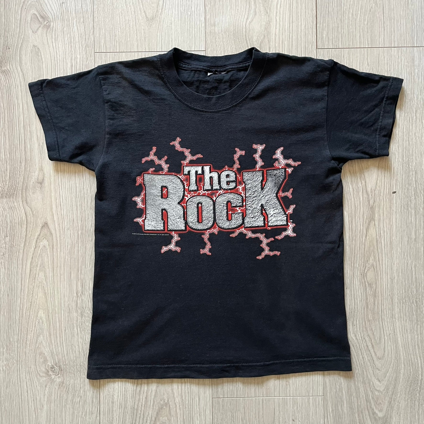 The Rock “Most Electrifying Man” 2000 Tee is a vintage WWE shirt featuring bold metallic-textured lettering with red lightning accents. The front proudly displays “The Rock,” while the back proclaims “The Most Electrifying Man in Sports Entertainment.” Licensed by World Wrestling Federation (WWF), this 2000 release is a must-have collectible for wrestling fans!