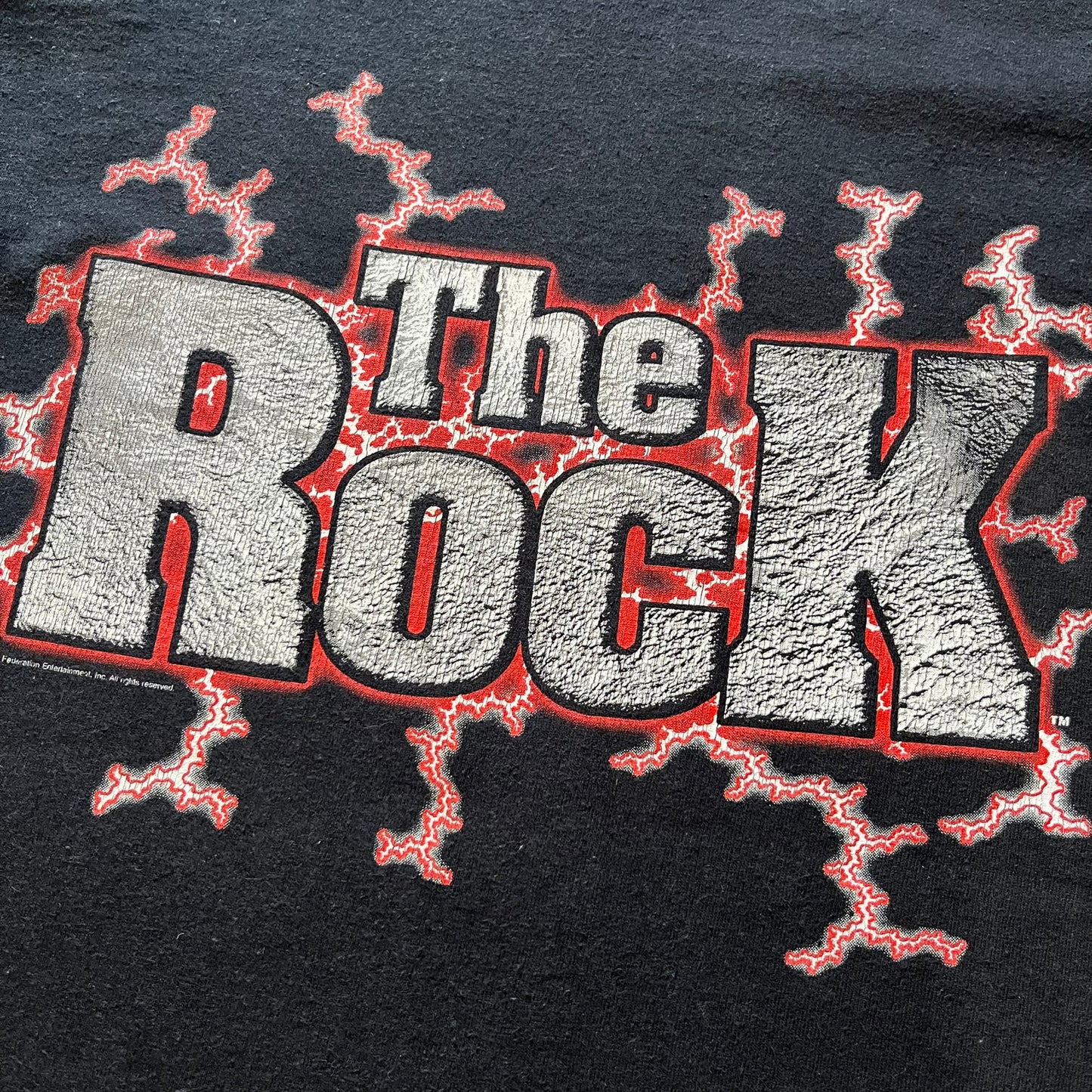 The Rock “Most Electrifying Man” 2000 Tee is a vintage WWE shirt featuring bold metallic-textured lettering with red lightning accents. The front proudly displays “The Rock,” while the back proclaims “The Most Electrifying Man in Sports Entertainment.” Licensed by World Wrestling Federation (WWF), this 2000 release is a must-have collectible for wrestling fans!