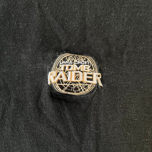Tomb Raider 2001 Embroidered Logo Tee is a minimalistic black, 100% cotton T-shirt with an embroidered Lara Croft Tomb Raider logo in white and gold tones at the center. The neck tag carries 2001 Paramount licensing.