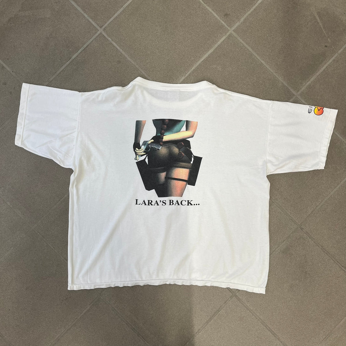 Tomb Raider III 1998 “Lara’s Back” White Tee is a boxy white T-shirt made on a Sierra Teez blank, with a stone-styled Tomb Raider III: Adventures of Lara Croft logo on the front. The back has a low-poly graphic of Lara Croft ass with “Lara’s Back...” text underneath, and the sleeve carries Eidos and Core logos.