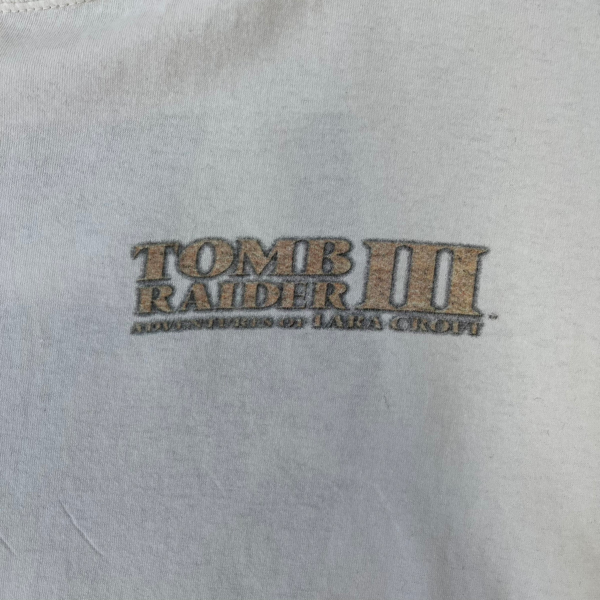 Tomb Raider III 1998 “Lara’s Back” White Tee is a boxy white T-shirt made on a Sierra Teez blank, with a stone-styled Tomb Raider III: Adventures of Lara Croft logo on the front. The back has a low-poly graphic of Lara Croft ass with “Lara’s Back...” text underneath, and the sleeve carries Eidos and Core logos.
