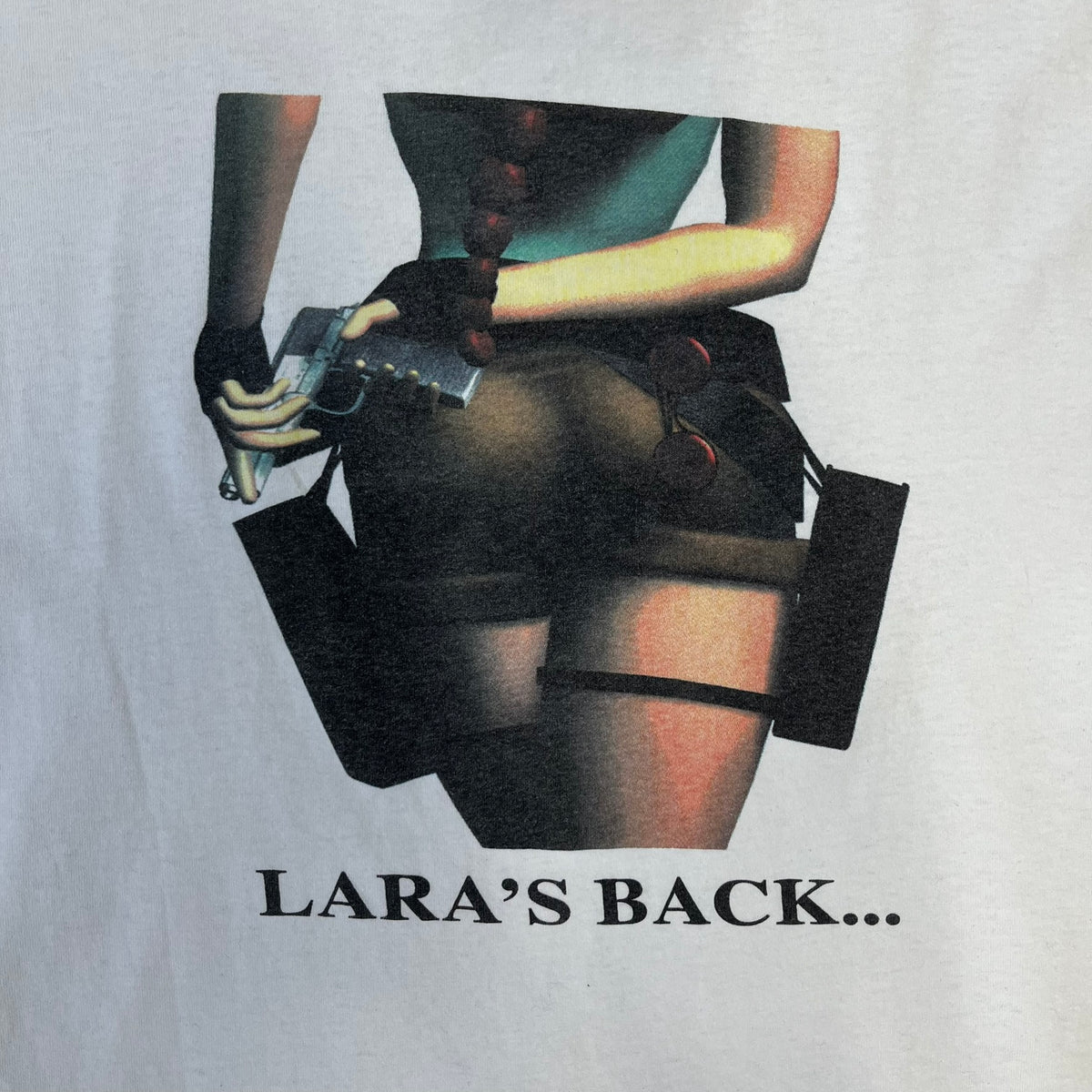 Tomb Raider III 1998 “Lara’s Back” White Tee is a boxy white T-shirt made on a Sierra Teez blank, with a stone-styled Tomb Raider III: Adventures of Lara Croft logo on the front. The back has a low-poly graphic of Lara Croft ass with “Lara’s Back...” text underneath, and the sleeve carries Eidos and Core logos.