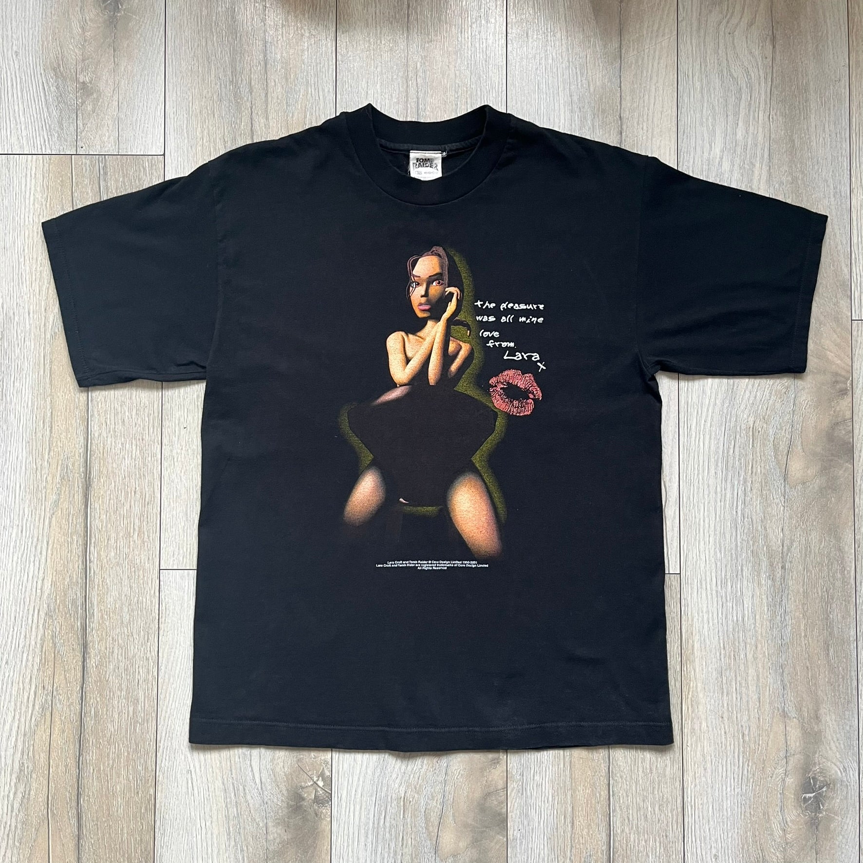 Tomb Raider Lara Croft 2001 Kiss Tee is a black T-shirt made in Spain from 100% cotton. The front features a naked low-poly game version of Lara Croft kneeling with a green glow background, next to a handwritten-style text reading “The pleasure was all mine, love from Lara x” and a glitter red kiss graphic. Small white licensing text reads Core Design Limited 1996–2001, and the neck tag includes Tomb Raider, Core, and Eidos branding.