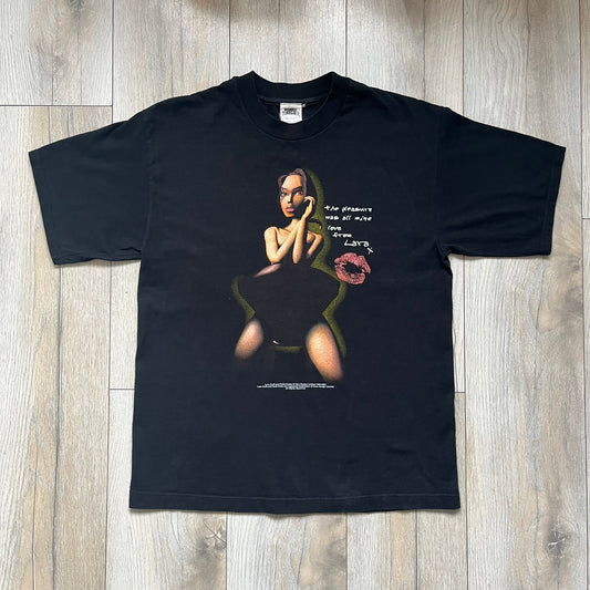 Tomb Raider Lara Croft 2001 Kiss Tee is a black T-shirt made in Spain from 100% cotton. The front features a naked low-poly game version of Lara Croft kneeling with a green glow background, next to a handwritten-style text reading “The pleasure was all mine, love from Lara x” and a glitter red kiss graphic. Small white licensing text reads Core Design Limited 1996–2001, and the neck tag includes Tomb Raider, Core, and Eidos branding.