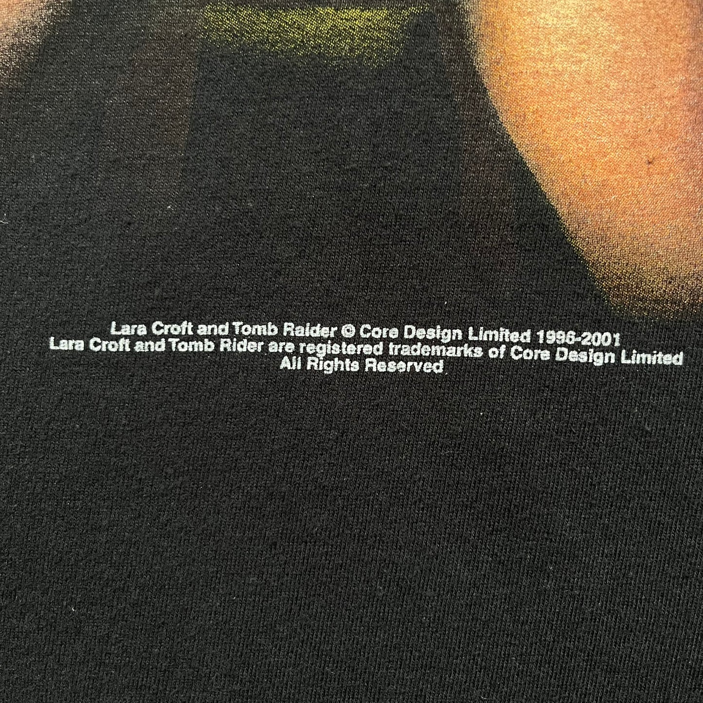 Tomb Raider Lara Croft 2001 Kiss Tee is a black T-shirt made in Spain from 100% cotton. The front features a naked low-poly game version of Lara Croft kneeling with a green glow background, next to a handwritten-style text reading “The pleasure was all mine, love from Lara x” and a glitter red kiss graphic. Small white licensing text reads Core Design Limited 1996–2001, and the neck tag includes Tomb Raider, Core, and Eidos branding.