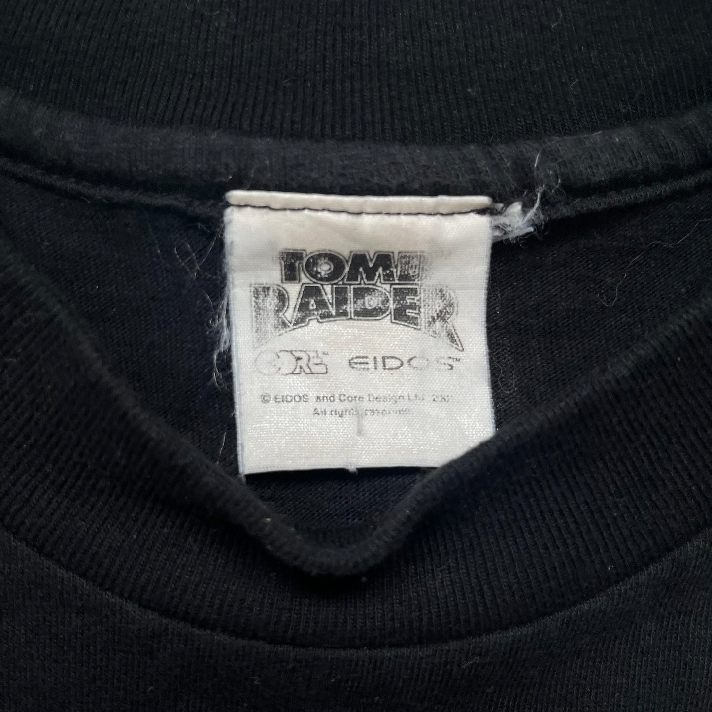Tomb Raider Lara Croft 2001 Kiss Tee is a black T-shirt made in Spain from 100% cotton. The front features a naked low-poly game version of Lara Croft kneeling with a green glow background, next to a handwritten-style text reading “The pleasure was all mine, love from Lara x” and a glitter red kiss graphic. Small white licensing text reads Core Design Limited 1996–2001, and the neck tag includes Tomb Raider, Core, and Eidos branding.