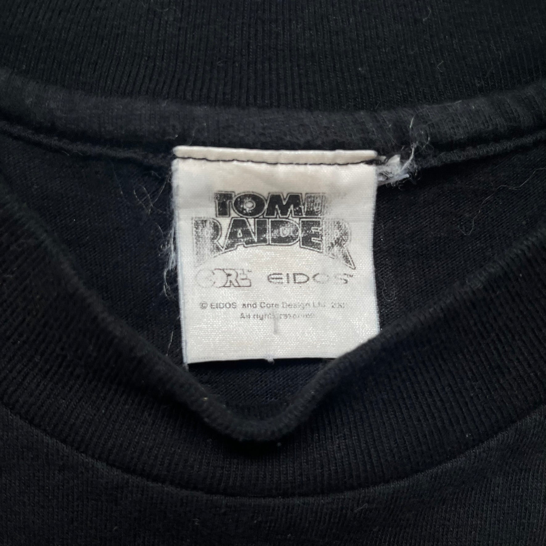 Tomb Raider Lara Croft 2001 Kiss Tee is a black T-shirt made in Spain from 100% cotton. The front features a naked low-poly game version of Lara Croft kneeling with a green glow background, next to a handwritten-style text reading “The pleasure was all mine, love from Lara x” and a glitter red kiss graphic. Small white licensing text reads Core Design Limited 1996–2001, and the neck tag includes Tomb Raider, Core, and Eidos branding.