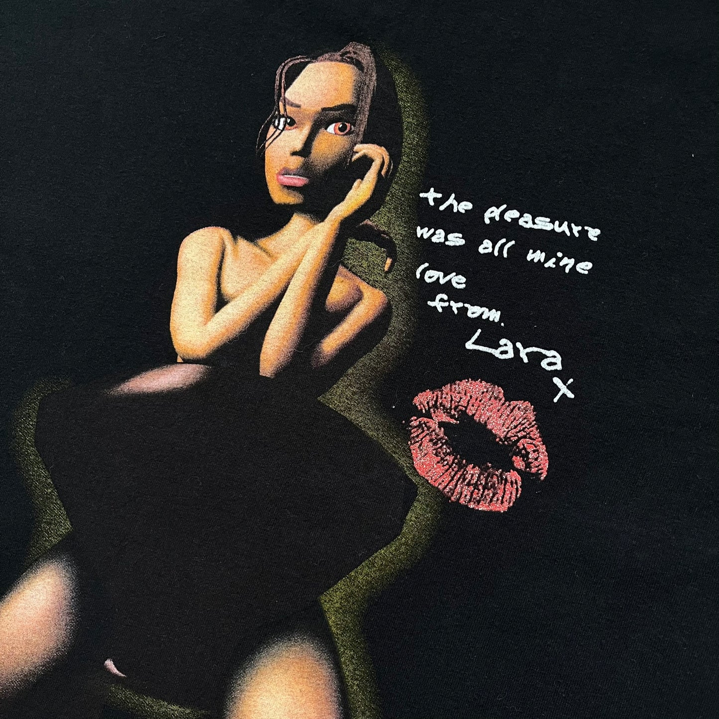 Tomb Raider Lara Croft 2001 Kiss Tee is a black T-shirt made in Spain from 100% cotton. The front features a naked low-poly game version of Lara Croft kneeling with a green glow background, next to a handwritten-style text reading “The pleasure was all mine, love from Lara x” and a glitter red kiss graphic. Small white licensing text reads Core Design Limited 1996–2001, and the neck tag includes Tomb Raider, Core, and Eidos branding.