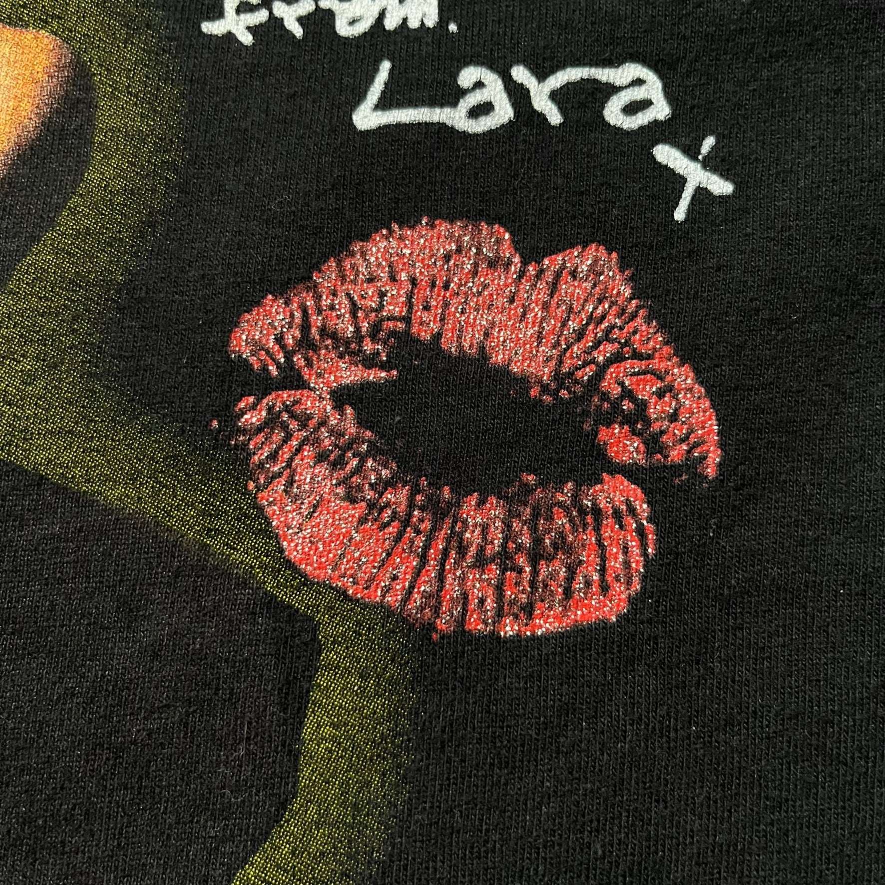 Tomb Raider Lara Croft 2001 Kiss Tee is a black T-shirt made in Spain from 100% cotton. The front features a naked low-poly game version of Lara Croft kneeling with a green glow background, next to a handwritten-style text reading “The pleasure was all mine, love from Lara x” and a glitter red kiss graphic. Small white licensing text reads Core Design Limited 1996–2001, and the neck tag includes Tomb Raider, Core, and Eidos branding.
