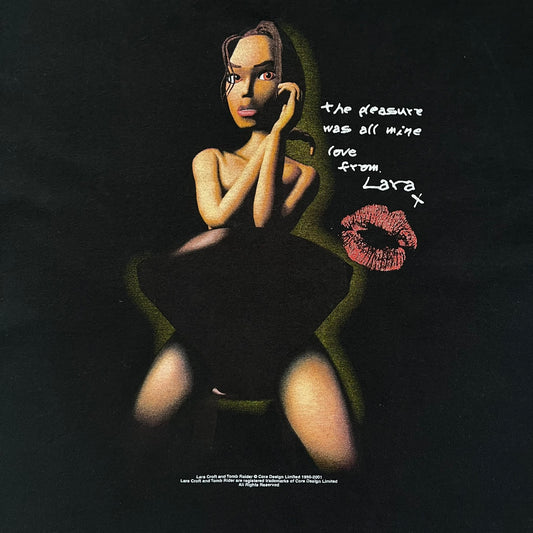 Tomb Raider Lara Croft 2001 Kiss Tee is a black T-shirt made in Spain from 100% cotton. The front features a naked low-poly game version of Lara Croft kneeling with a green glow background, next to a handwritten-style text reading “The pleasure was all mine, love from Lara x” and a glitter red kiss graphic. Small white licensing text reads Core Design Limited 1996–2001, and the neck tag includes Tomb Raider, Core, and Eidos branding.