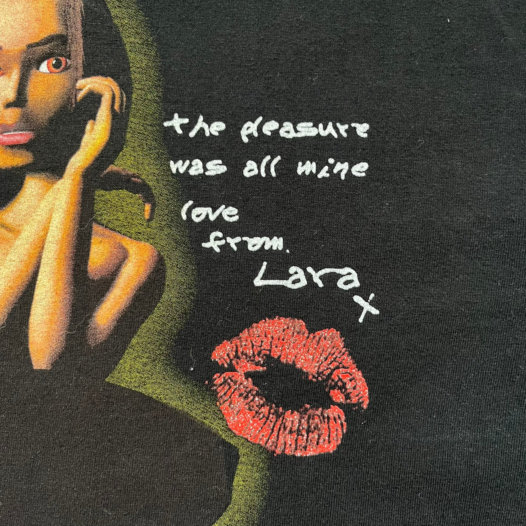 Tomb Raider Lara Croft 2001 Kiss Tee is a black T-shirt made in Spain from 100% cotton. The front features a naked low-poly game version of Lara Croft kneeling with a green glow background, next to a handwritten-style text reading “The pleasure was all mine, love from Lara x” and a glitter red kiss graphic. Small white licensing text reads Core Design Limited 1996–2001, and the neck tag includes Tomb Raider, Core, and Eidos branding.