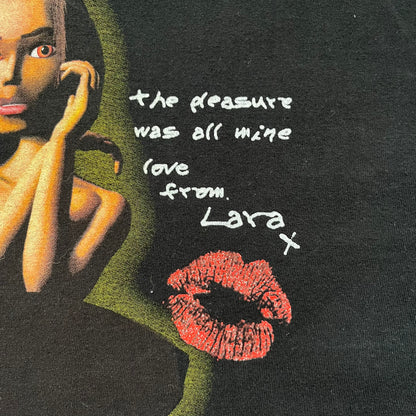 Tomb Raider Lara Croft 2001 Kiss Tee is a black T-shirt made in Spain from 100% cotton. The front features a naked low-poly game version of Lara Croft kneeling with a green glow background, next to a handwritten-style text reading “The pleasure was all mine, love from Lara x” and a glitter red kiss graphic. Small white licensing text reads Core Design Limited 1996–2001, and the neck tag includes Tomb Raider, Core, and Eidos branding.