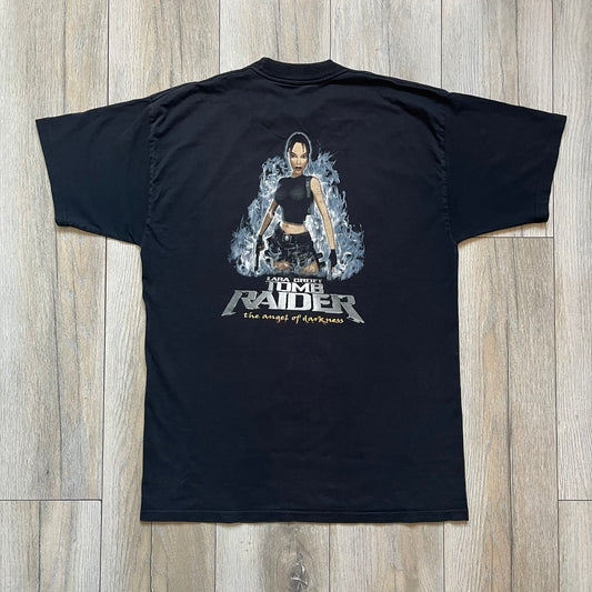 Tomb Raider: The Angel of Darkness 2003 Tee is a black washed T-shirt featuring a small game logo on the front chest. The back displays a large graphic based on the game cover, with Lara Croft in front of a flame rendered in early 3D-style artwork, with the game title printed below.
