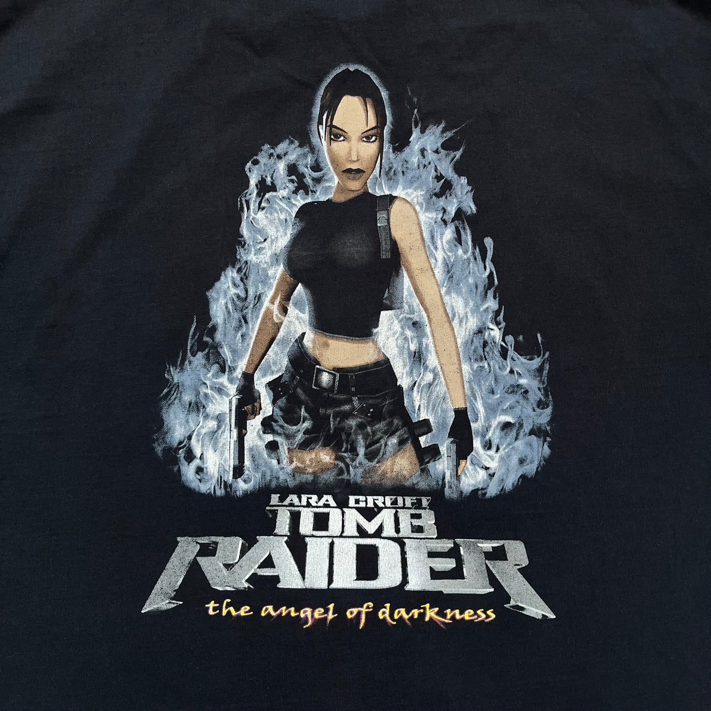 Tomb Raider: The Angel of Darkness 2003 Tee is a black washed T-shirt featuring a small game logo on the front chest. The back displays a large graphic based on the game cover, with Lara Croft in front of a flame rendered in early 3D-style artwork, with the game title printed below.

