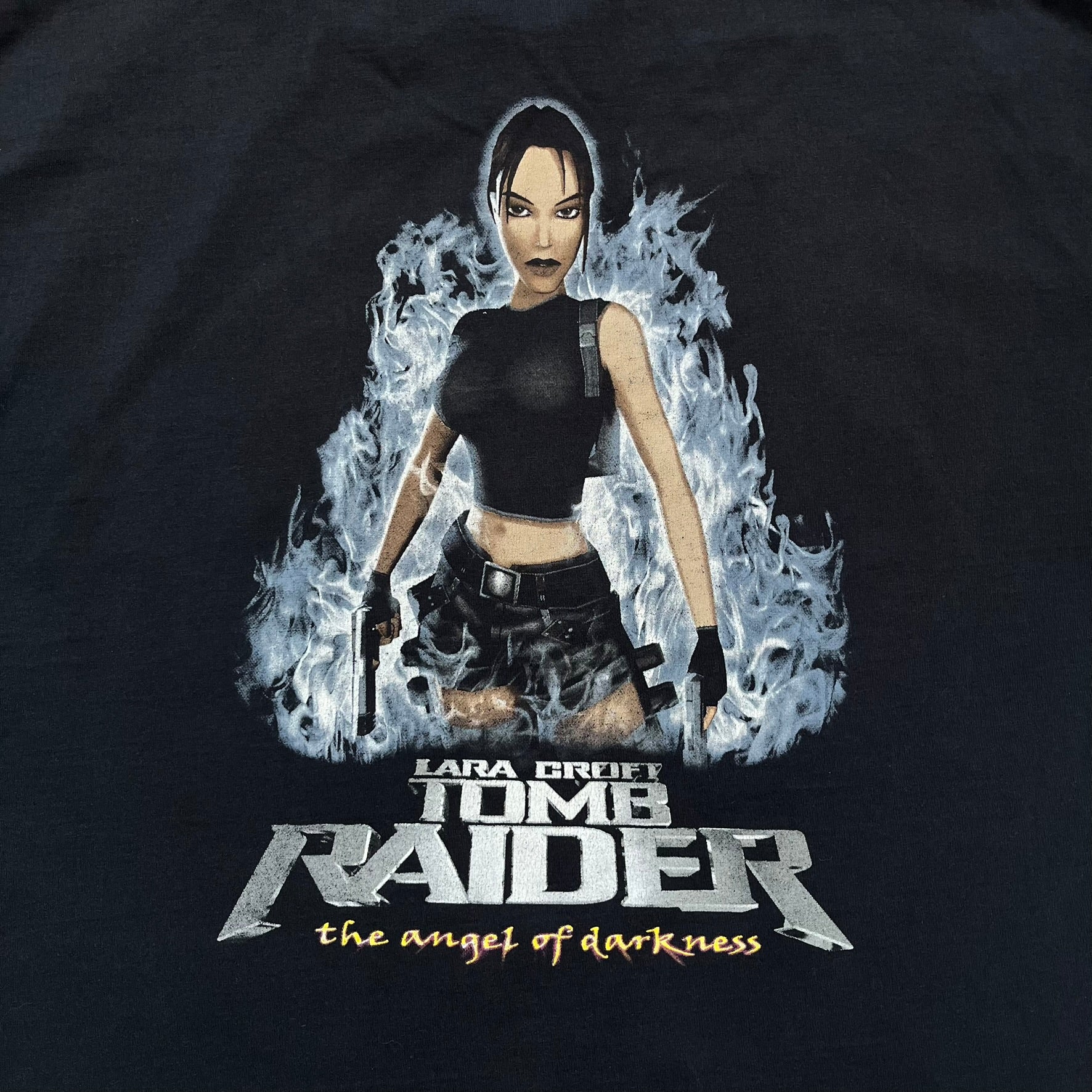 Tomb Raider: The Angel of Darkness 2003 Tee is a black washed T-shirt featuring a small game logo on the front chest. The back displays a large graphic based on the game cover, with Lara Croft in front of a flame rendered in early 3D-style artwork, with the game title printed below.
