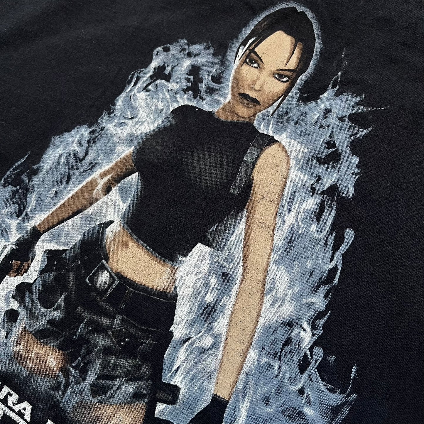 Tomb Raider: The Angel of Darkness 2003 Tee is a black washed T-shirt featuring a small game logo on the front chest. The back displays a large graphic based on the game cover, with Lara Croft in front of a flame rendered in early 3D-style artwork, with the game title printed below.
