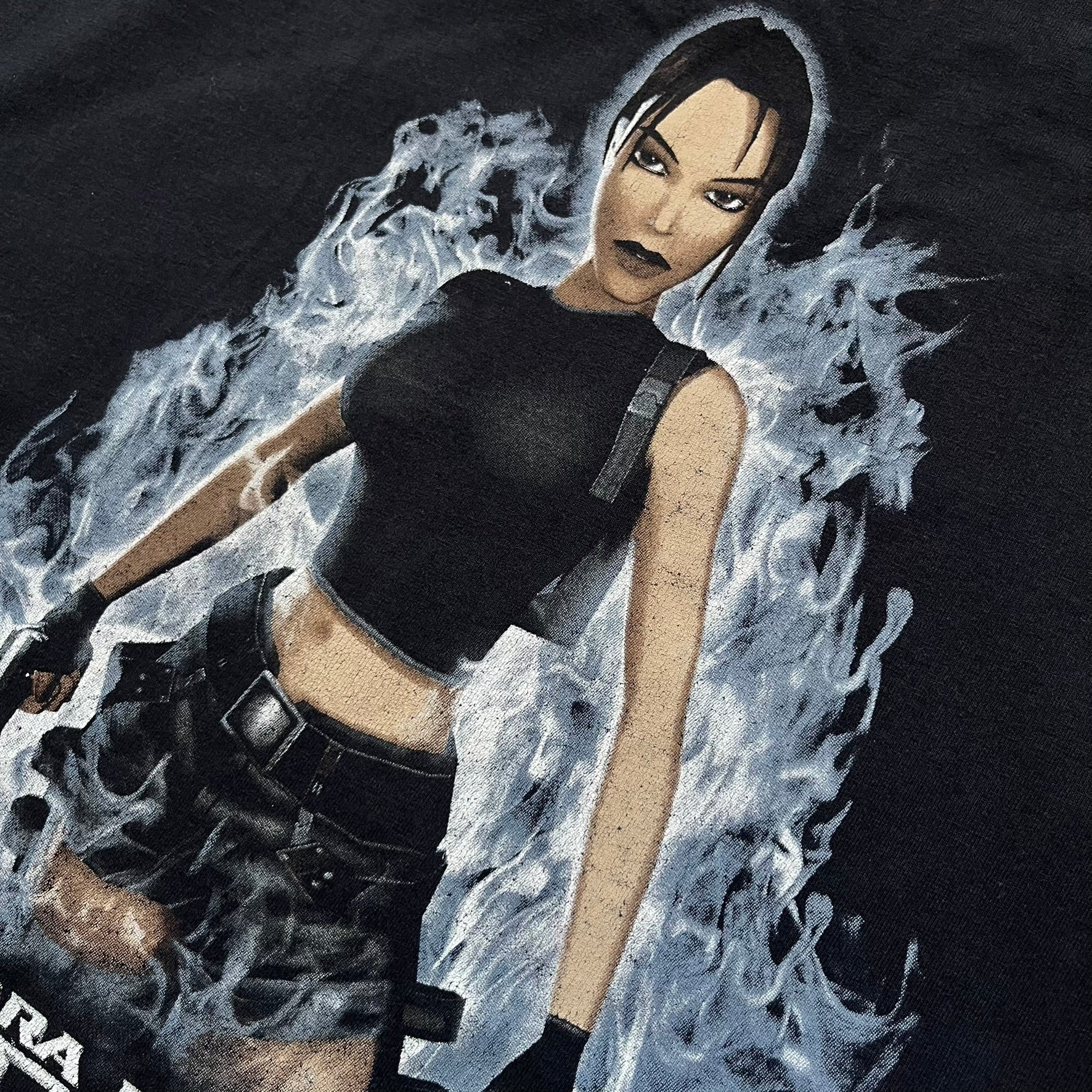 Tomb Raider: The Angel of Darkness 2003 Tee is a black washed T-shirt featuring a small game logo on the front chest. The back displays a large graphic based on the game cover, with Lara Croft in front of a flame rendered in early 3D-style artwork, with the game title printed below.
