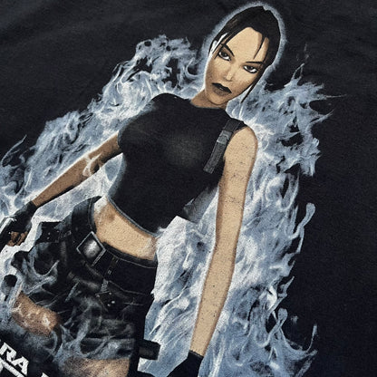 Tomb Raider: The Angel of Darkness 2003 Tee is a black washed T-shirt featuring a small game logo on the front chest. The back displays a large graphic based on the game cover, with Lara Croft in front of a flame rendered in early 3D-style artwork, with the game title printed below.
