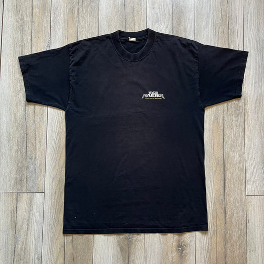 Tomb Raider: The Angel of Darkness 2003 Tee is a black washed T-shirt featuring a small game logo on the front chest. The back displays a large graphic based on the game cover, with Lara Croft in front of a flame rendered in early 3D-style artwork, with the game title printed below.
