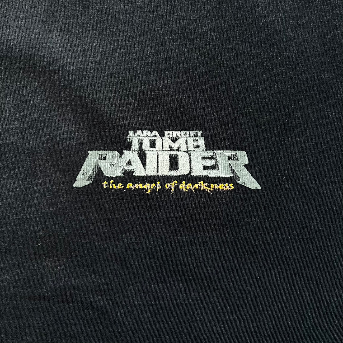 Tomb Raider: The Angel of Darkness 2003 Tee is a black washed T-shirt featuring a small game logo on the front chest. The back displays a large graphic based on the game cover, with Lara Croft in front of a flame rendered in early 3D-style artwork, with the game title printed below.
