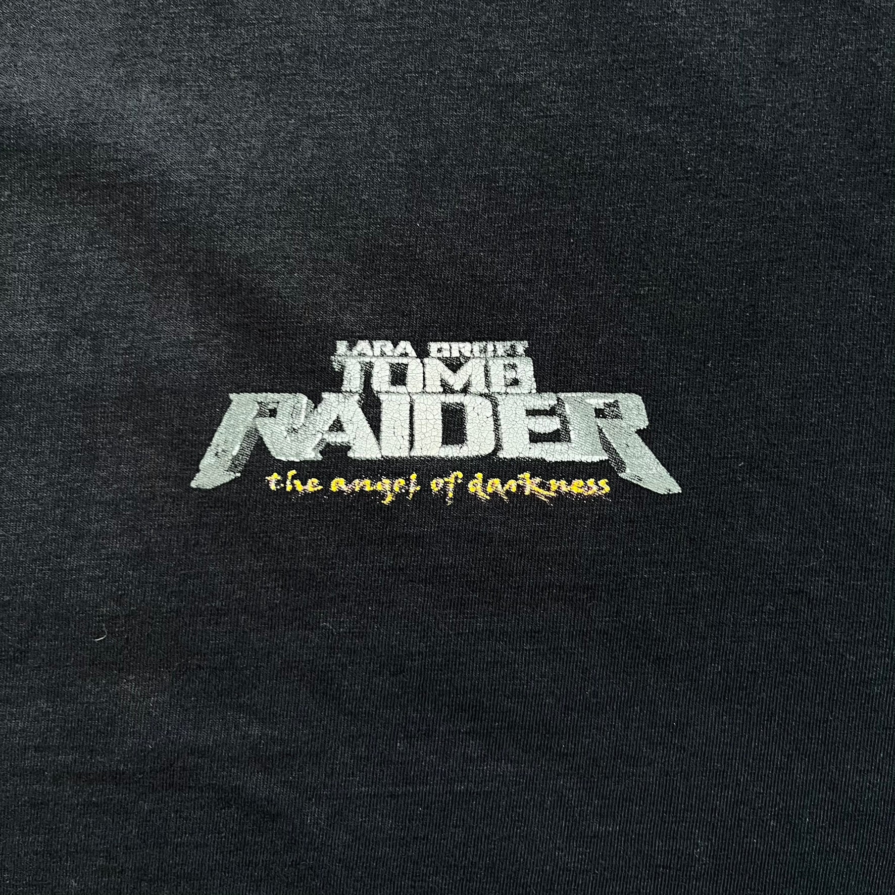 Tomb Raider: The Angel of Darkness 2003 Tee is a black washed T-shirt featuring a small game logo on the front chest. The back displays a large graphic based on the game cover, with Lara Croft in front of a flame rendered in early 3D-style artwork, with the game title printed below.
