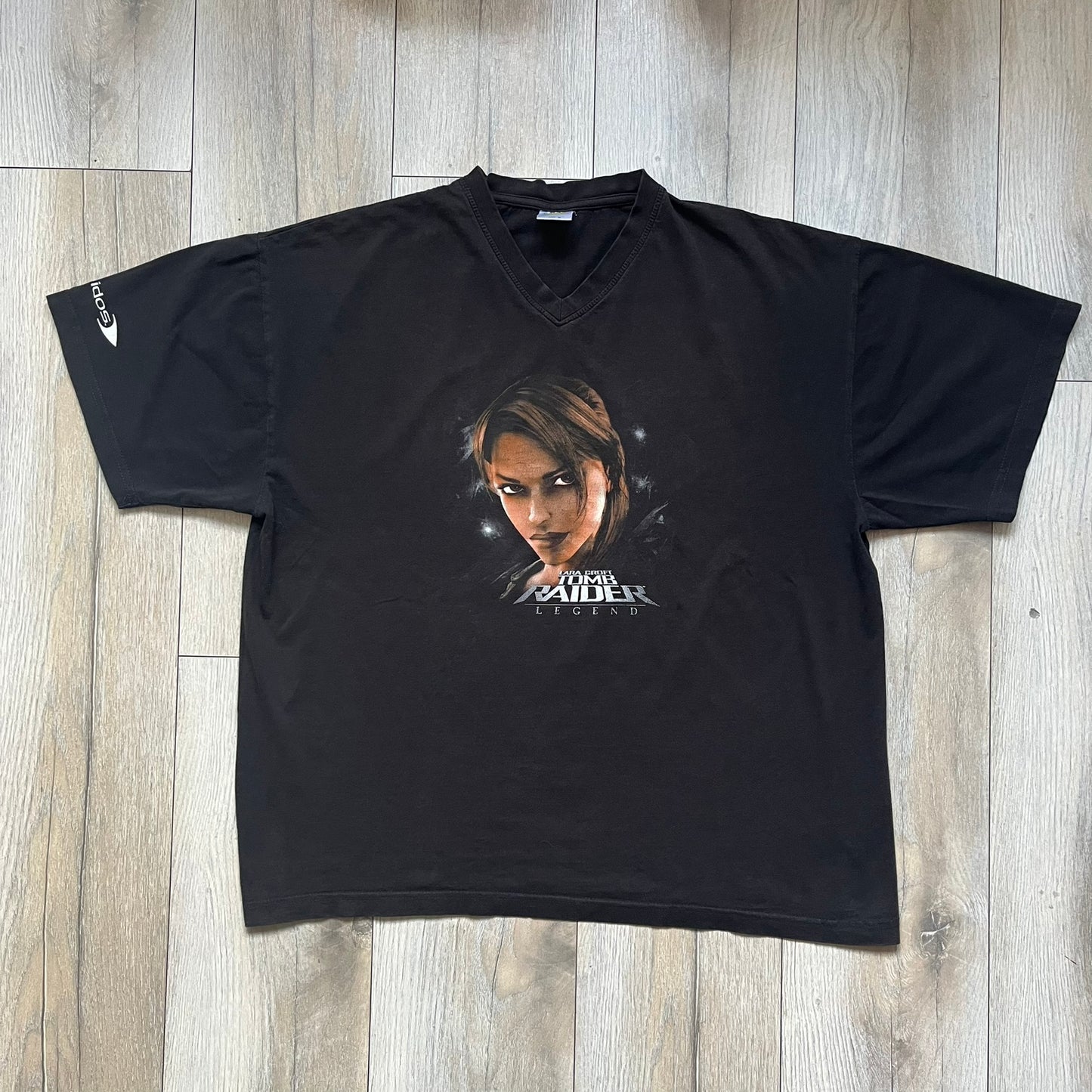 Tomb Raider Lara Croft 2006 Legend Tee is a black V-neck T-shirt made from 100% cotton on a Hanes blank. The front features the Lara Croft: Tomb Raider Legend cover graphic with Lara Croft’s face and the game title printed below. A white Eidos logo is printed on the sleeve, referencing the official game publisher.