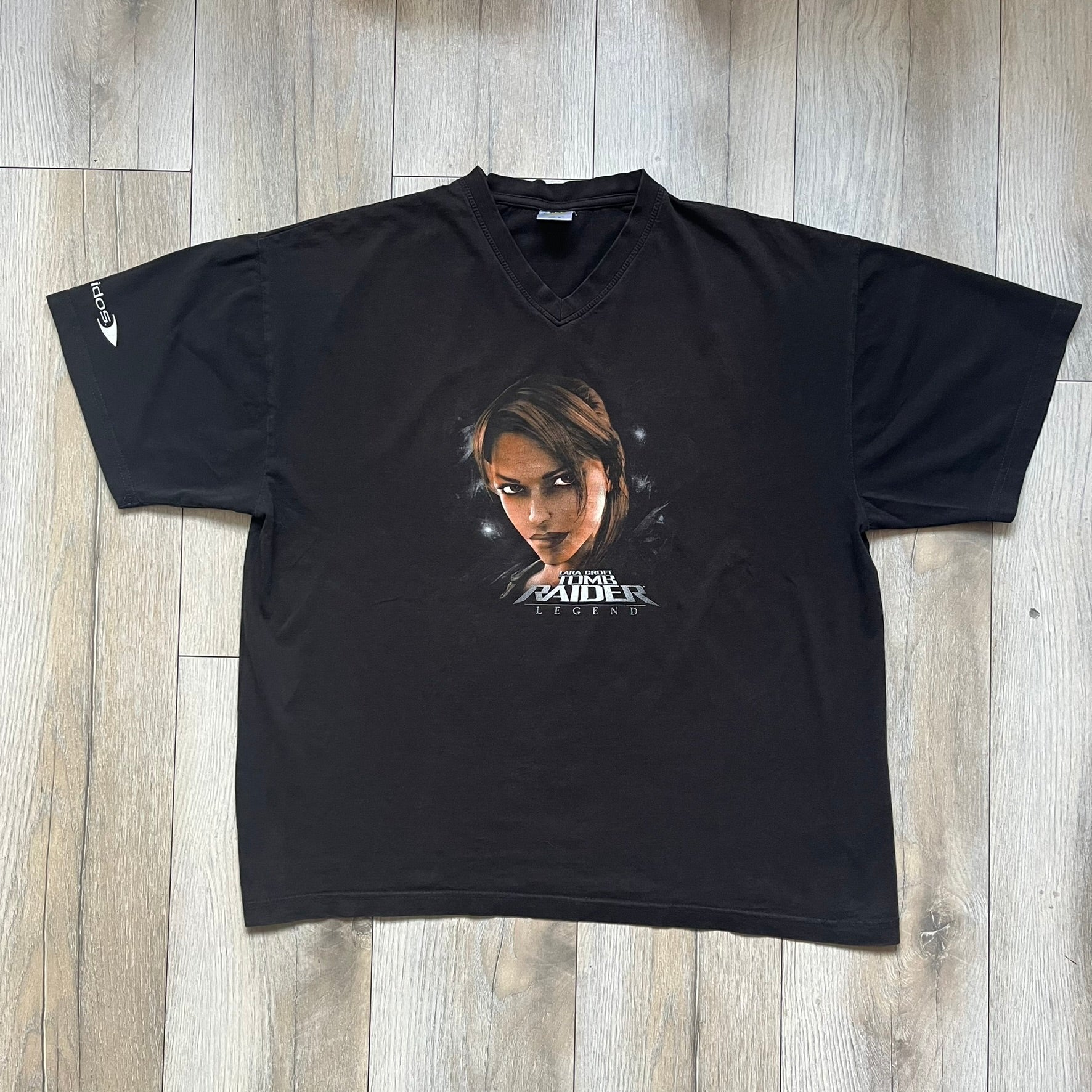Tomb Raider Lara Croft 2006 Legend Tee is a black V-neck T-shirt made from 100% cotton on a Hanes blank. The front features the Lara Croft: Tomb Raider Legend cover graphic with Lara Croft’s face and the game title printed below. A white Eidos logo is printed on the sleeve, referencing the official game publisher.