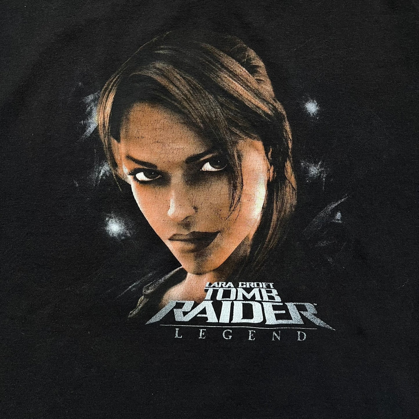 Tomb Raider Lara Croft 2006 Legend Tee is a black V-neck T-shirt made from 100% cotton on a Hanes blank. The front features the Lara Croft: Tomb Raider Legend cover graphic with Lara Croft’s face and the game title printed below. A white Eidos logo is printed on the sleeve, referencing the official game publisher.
