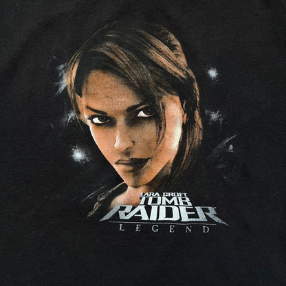 Tomb Raider Lara Croft 2006 Legend Tee is a black V-neck T-shirt made from 100% cotton on a Hanes blank. The front features the Lara Croft: Tomb Raider Legend cover graphic with Lara Croft’s face and the game title printed below. A white Eidos logo is printed on the sleeve, referencing the official game publisher.