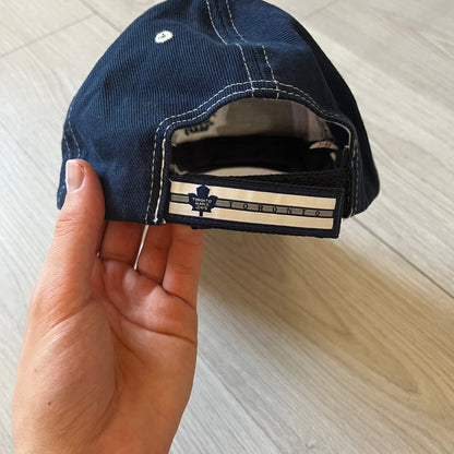 Hand holding a navy blue cap with a brand logo on a light wooden surface