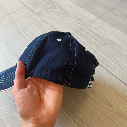 Hand holding a navy blue cap with white stitching on a light wooden floor.