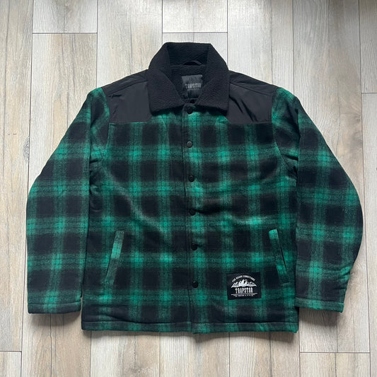 The Trapstar Irongate Green Plaid Lumberjack Jacket is a woven jacket in a green and black checked pattern, made from a 95% polyester and 5% wool outer with a full polyester lining. It features contrast shell panels at the shoulders, exposed popper fastenings at the front, a classic collar, and adjustable popper tabs at the cuffs. Details include side slip pockets, a Trapstar brand patch at the front hem, and a large Trapstar logo embroidered across the upper back yoke.