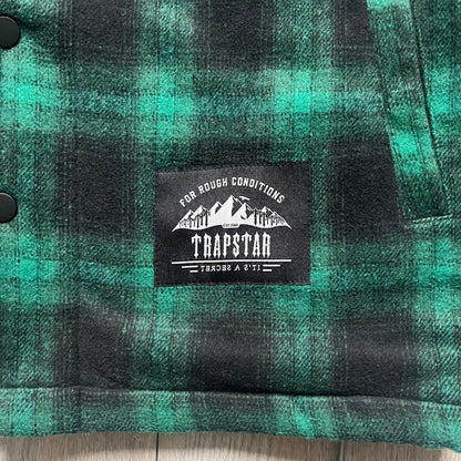 The Trapstar Irongate Green Plaid Lumberjack Jacket is a woven jacket in a green and black checked pattern, made from a 95% polyester and 5% wool outer with a full polyester lining. It features contrast shell panels at the shoulders, exposed popper fastenings at the front, a classic collar, and adjustable popper tabs at the cuffs. Details include side slip pockets, a Trapstar brand patch at the front hem, and a large Trapstar logo embroidered across the upper back yoke.
