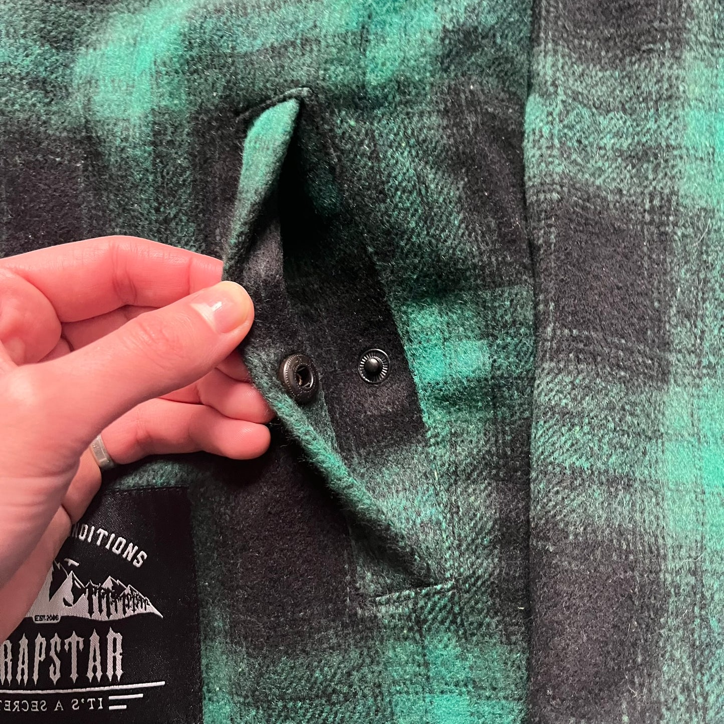 The Trapstar Irongate Green Plaid Lumberjack Jacket is a woven jacket in a green and black checked pattern, made from a 95% polyester and 5% wool outer with a full polyester lining. It features contrast shell panels at the shoulders, exposed popper fastenings at the front, a classic collar, and adjustable popper tabs at the cuffs. Details include side slip pockets, a Trapstar brand patch at the front hem, and a large Trapstar logo embroidered across the upper back yoke.